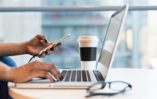 Photo of a person using their mobile smartphone while at a computer to illustrate the launch of OpenAI Operator that promises to democratise agentic AI by bringing agents into the browser.